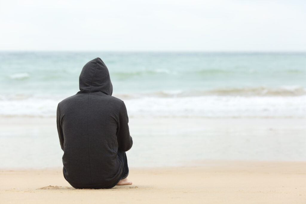 Rear view portrait of one teenager boy thinking alone and watching the sea sitting on the sand of the beach with the horizon in the background